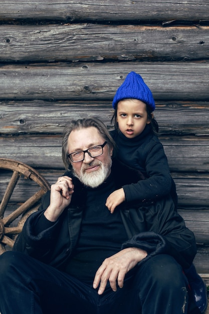 Petit-fils dans un chapeau bleu et des bottes avec un grand-père dans un portemanteau en peau de mouton dans une maison en bois remise dans le village au printemps en Russie