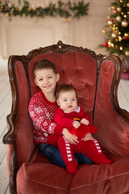 Petit frère embrasse sa petite sœur dans une chaise rétro rouge près de l'arbre de Noël