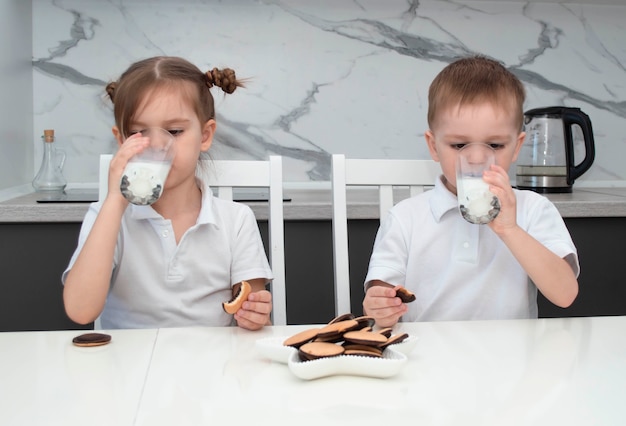 Le petit frère et la soeur blonds heureux et mignons rient et mangent des biscuits assis à table.