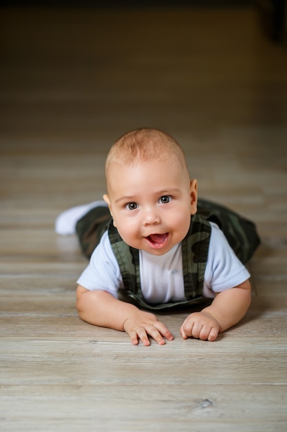 Petit garçon de 8 mois en salopette, chemise blanche et chaussettes blanches rampant sur le sol et souriant