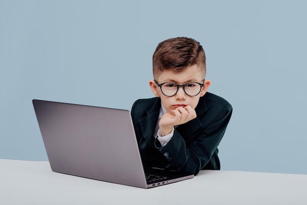 Petit garçon d'affaires bouleversé avec des lunettes à l'aide d'un petit patron d'ordinateur portable dans un enfant drôle de bureau isolé sur bl...