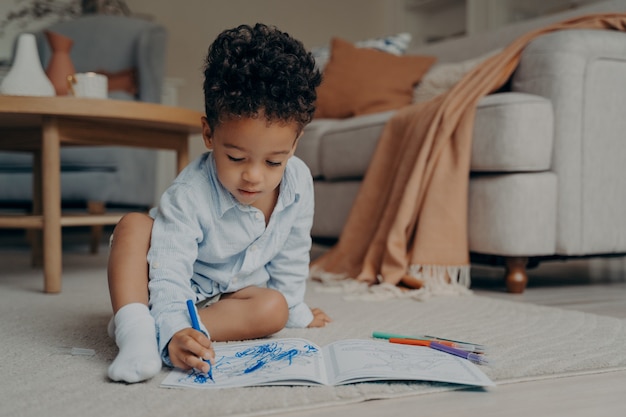 Petit garçon africain assis sur le sol et dessinant avec un stylo-feutre bleu