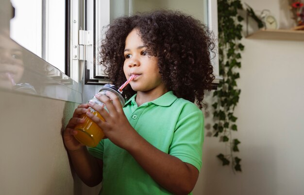 Un petit garçon afro boit du jus avec de la paille à la maison à la fenêtre