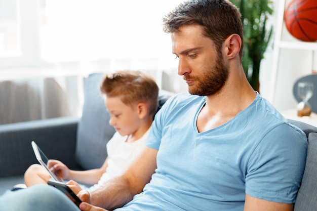 Photo petit garçon à l'aide de tablette numérique et son père à l'aide de smartphone s'asseoir ensemble sur le canapé à la maison