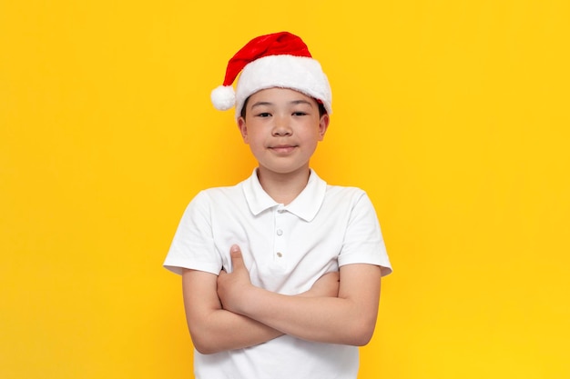 petit garçon asiatique en bonnet de noel se tient avec les bras croisés sur fond jaune isolé