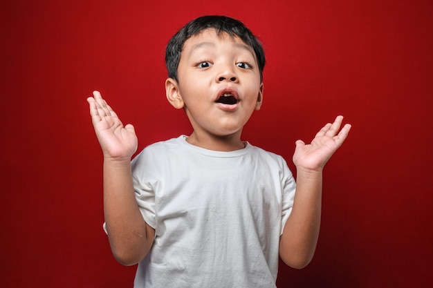 Petit garçon asiatique choqué en t-shirt blanc touchant son visage sur fond rouge