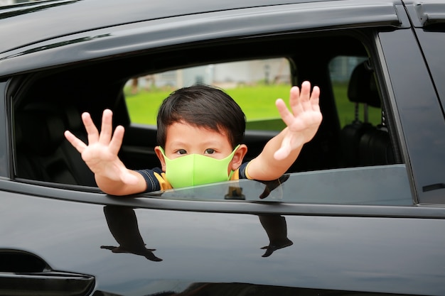 Un petit garçon asiatique portant un masque d'hygiène sort la tête de la fenêtre de la voiture avec un geste de signe d'arrêt de la main pendant l'épidémie de coronavirus (covid-19)