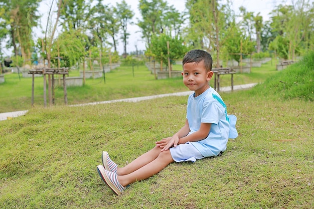 Photo petit garçon asiatique regardant la caméra et assis sur l'herbe verte dans le jardin