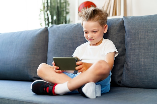 Petit garçon assis sur un canapé à la maison et à l'aide de smartphone