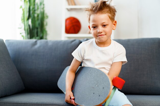 Petit garçon assis sur un canapé à la maison et tenant une planche à roulettes