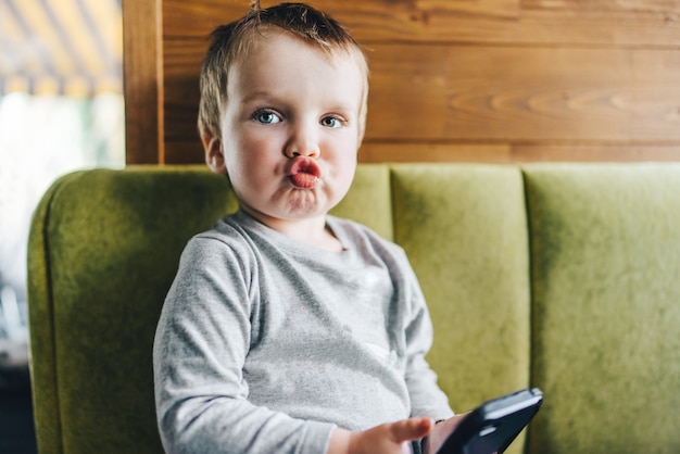 Petit garçon assis sur un canapé avec un téléphone portable dans les mains et envoyer un baiser