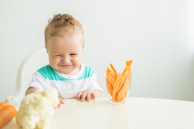 Petit garçon assis dans une chaise d'enfant mangeant des tranches de carottes sur fond blanc.