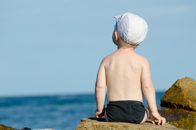 Petit garçon assis le dos à un rocher au bord de la mer en maillot de bain, ciel bleu, espace pour le texte