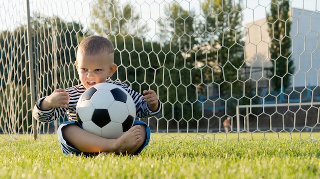 Petit garçon assis les jambes croisées sur le sol dans les poteaux avec un ballon de football en équilibre sur ses genoux