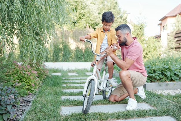 Petit garçon assis à vélo attendant que son père fixe la hauteur du guidon à l'extérieur par une journée d'été ensoleillée