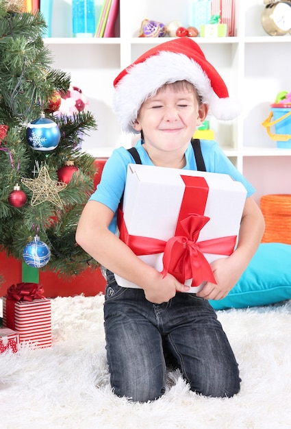 Le petit garçon au bonnet de Noel est assis près de l'arbre de Noël avec un cadeau dans les mains