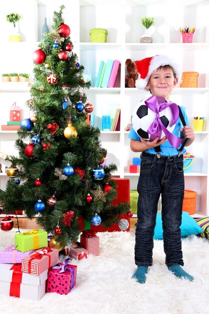 Le petit garçon au bonnet de Noel se tient près de l'arbre de Noël avec un ballon de football