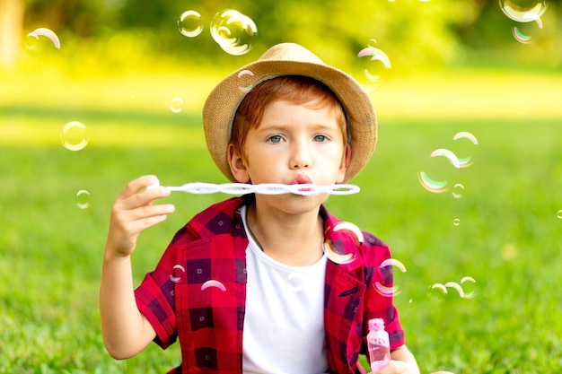 Un petit garçon aux cheveux roux gonfle des bulles de savon sur une pelouse verte dans un chapeau et une chemise rouge en été s'amusant à jouer