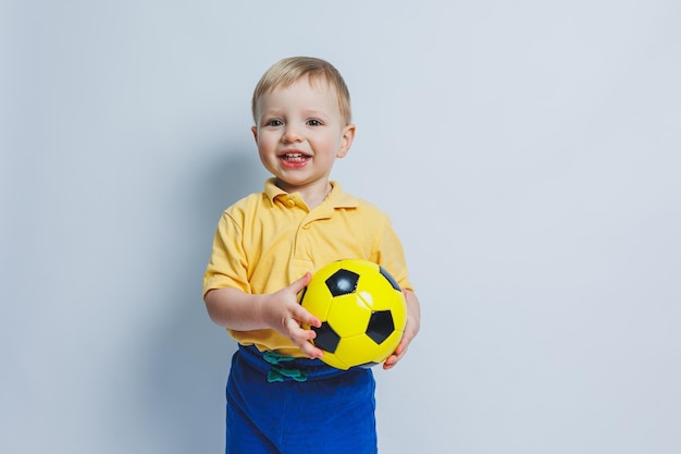 Un petit garçon avec un ballon de football dans ses mains sur un fond blanc un enfant est un joueur de football novice un sport pour les enfants Petit athlète Kit de football jaune et bleu pour les enfants