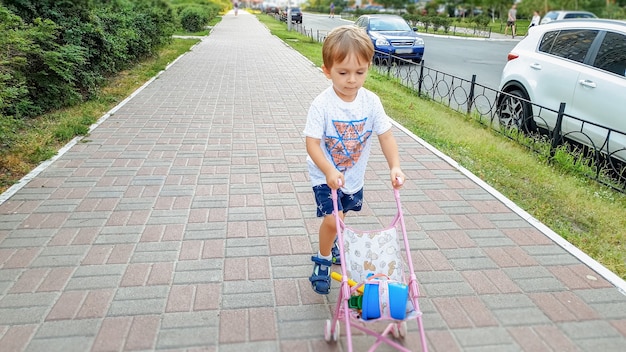 Petit garçon en bas âge marchant sur la route et poussant la poussette de jouet pour des poupées