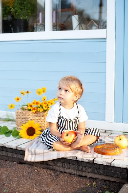 Petit garçon blond assis sur un porche en bois à la maison et mange une pomme un jour d'automne. l'enfant joue dans la cour à l'automne.