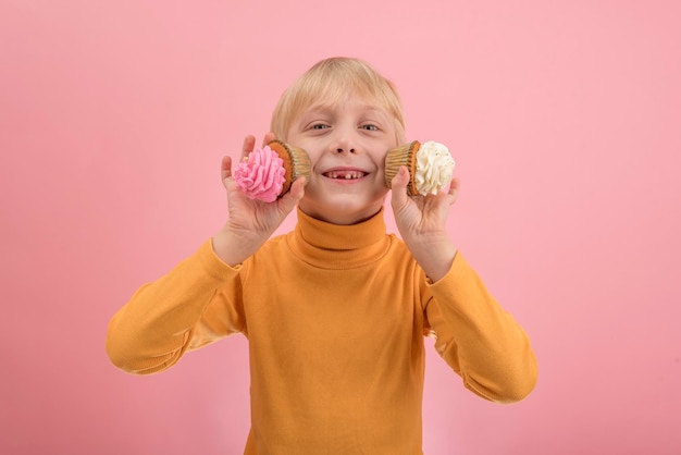 Petit garçon blond avec des cupcakes sur fond rose Garçon de 7 ans souriant avec des cupcakes à la crème blanche et rose