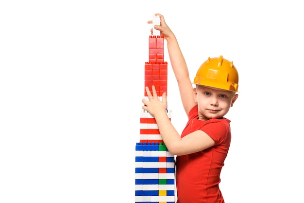 Photo petit garçon blond dans le casque de construction et une chemise rouge debout près de la tour construite à partir de concepteur de pièces