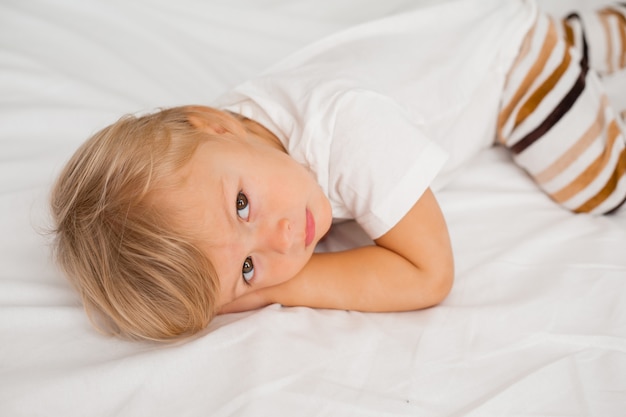 petit garçon blond dans un T-shirt blanc est allongé sur le ventre sur un drap blanc dans le lit