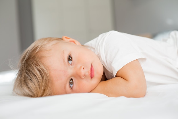 petit garçon blond dans un T-shirt blanc est allongé sur le ventre sur un drap blanc dans le lit