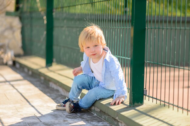 Petit garçon blond est assis près de la clôture.