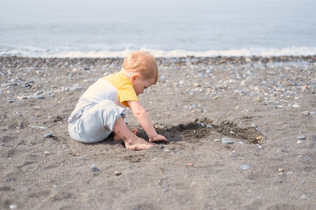 Petit garçon blond jouant avec des rochers et du sable sur la plage par une journée ensoleillée.