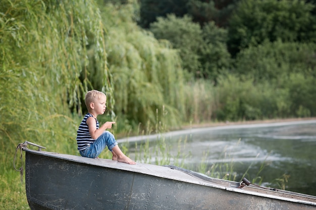Petit garçon blond joue au bord de la rivière avec une canne à pêche. herbe verte d'été et lac bleu.