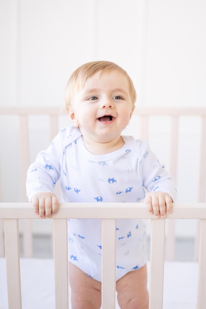 Un petit garçon blond souriant dans un berceau à la maison avec une literie en coton bleu et blanc se tient sur le côté et rit le matin le concept de produits et accessoires pour enfants