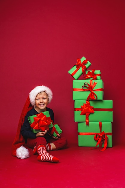 Petit garçon en bonnet de Noel est assis à côté de coffrets cadeaux de Noël pyramide sur rouge en Studio.