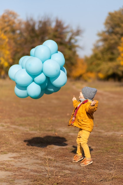 Photo un petit garçon avec une brassée de ballons se promène dans le parc en automne. arbres jaunes et boules bleues. enfant élégant.