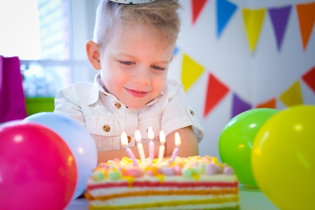 Petit garçon caucasien blond regardant des bougies sur un gâteau arc-en-ciel d'anniversaire faisant un vœu avant les coups