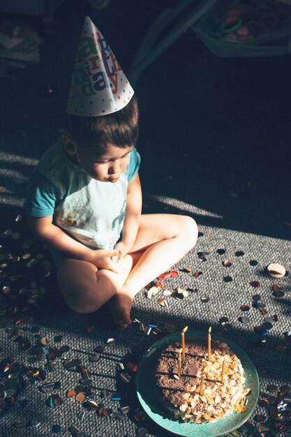 Petit garçon caucasien avec chapeau de fête souffle les lumières d'anniversaire assis sur le sol près de confettis