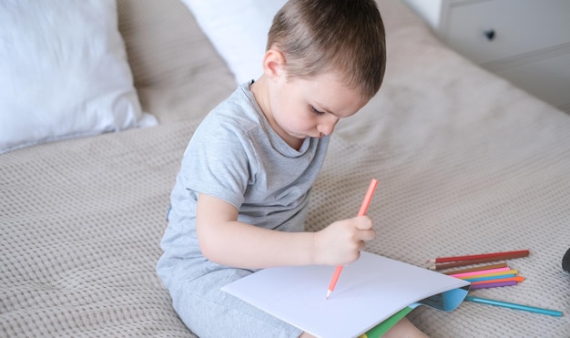 Un petit garçon caucasien dans un T-shirt gris et un short sur le lit dessine avec des crayons de couleur dans un croquis