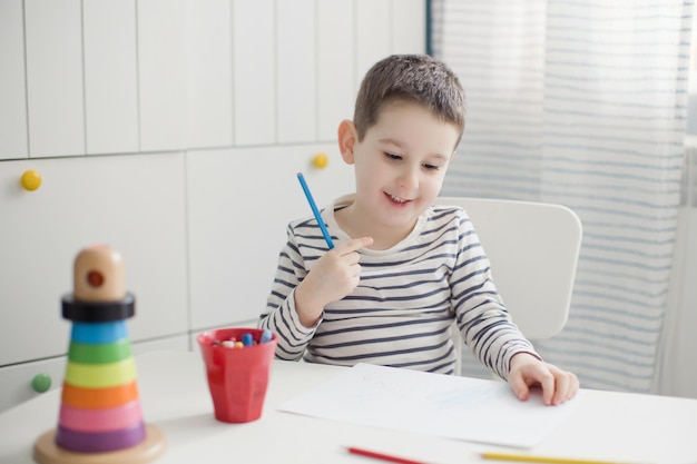 Petit garçon caucasien dessinant à la maison Enfant dans une pièce lumineuse assis sur une chaise blanche et dessinant sur un tableau blanc