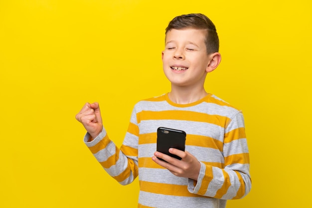 Petit garçon caucasien isolé sur fond jaune avec téléphone en position de victoire