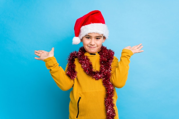 Petit garçon célébrant le jour de Noël portant un bonnet de Noel isolé fait échelle avec les bras, se sent heureux et confiant.