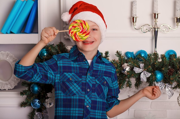Petit garçon en chapeau de père Noël tient une grosse sucette savoureuse dans sa main dans la chambre avec décoration de Noël