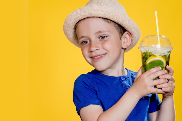 Petit garçon avec chapeau et t-shirt bleu tenant un cocktail mojito dans une tasse en plastique sur fond jaune