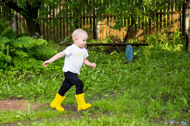Un petit garçon chauve en bottes jaunes court dans la campagne à travers des flaques d'eau à l'air frais