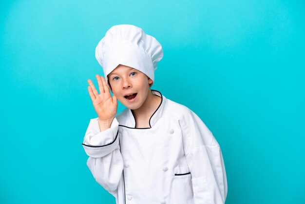 Petit garçon chef isolé sur fond bleu écoutant quelque chose en mettant la main sur l'oreille