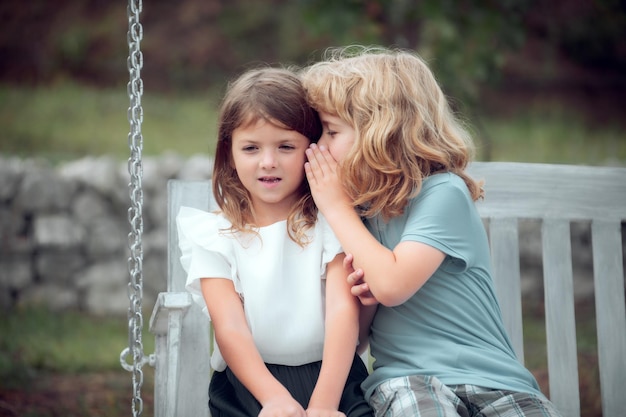 Petit garçon chuchote à la jolie fille dans l'oreille portrait de frère et soeur dans le parc d'été allumé à l'extérieur