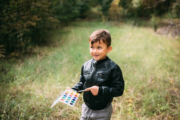 Un petit garçon de cinq ans se tient dans la forêt avec des peintures dans les mains, essayant de peindre les feuilles des arbres avec des peintures