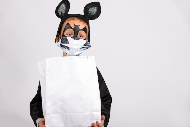 Petit garçon en costume de chauve-souris tenant un sac blanc de chocolats avec un espace vide. Enfant en masque médical sur mur gris.