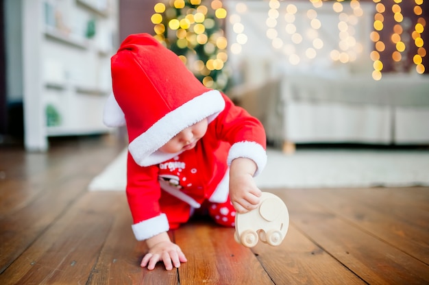 Petit garçon avec le costume du père Noël joue sur le sol avec une petite voiture en bois sur le fond des lumières du nouvel an