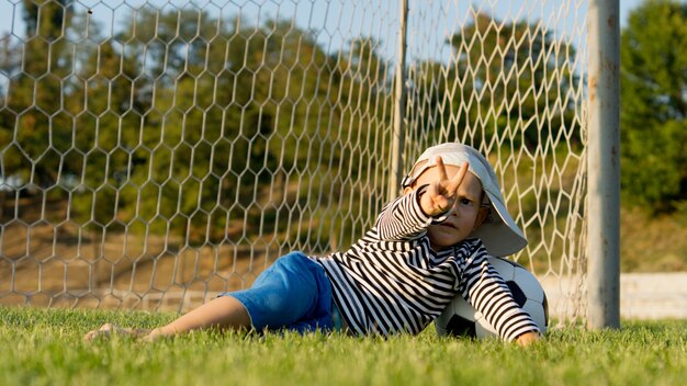 Petit garçon couché sur les poteaux sur un terrain de sport avec son ballon de football donnant un signe de victoire avec ses doigts